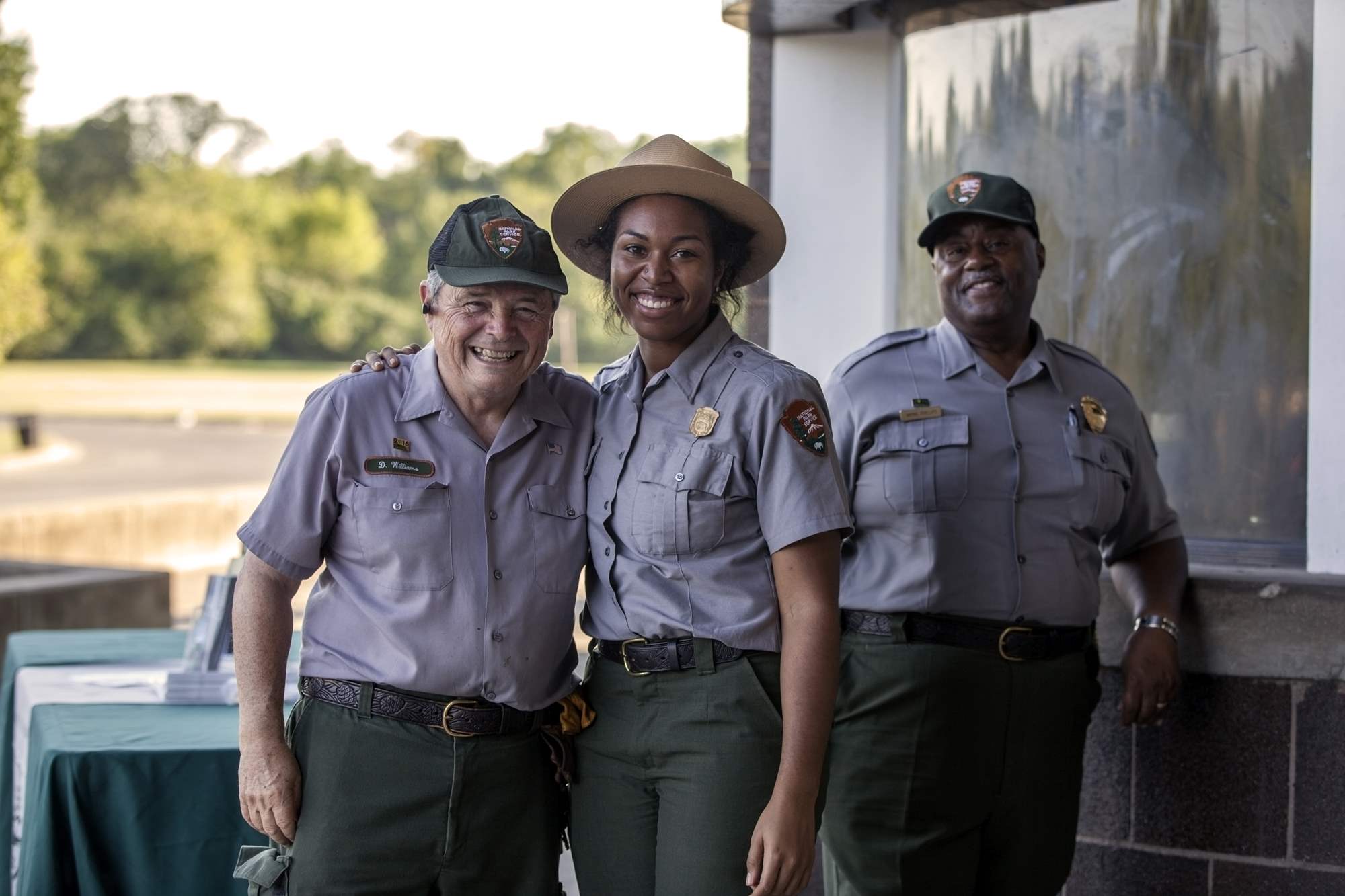 Three people stand in NPS uniforms. Only two wear badges. A woman wears a gold shield shaped badge and a man a large gold arrowhead shaped badge.